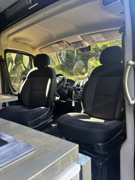 Interior of an RV rental: front seats & dashboard bathed in sunlight, with green trees visible through the windows.