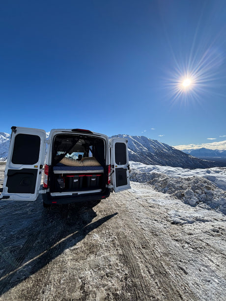 Enjoying mountain van life! A white RV rental van with open rear doors reveals a cozy bed, parked on a snowy road under a bright sun.