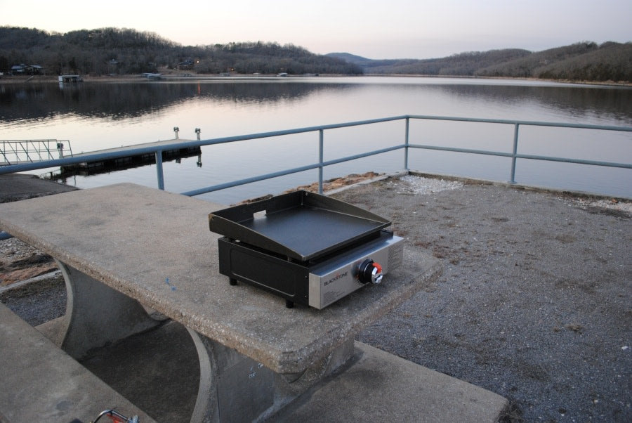A portable Blackstone griddle on a concrete picnic table by a calm lake with wooded hills, perfect for your next RV rental trip.