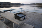 A portable Blackstone griddle on a concrete picnic table by a calm lake with wooded hills, perfect for your next RV rental trip.