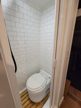 RV rental wet bath featuring white subway tile walls, a white toilet, and a slatted wood shower floor. Functional and compact design.