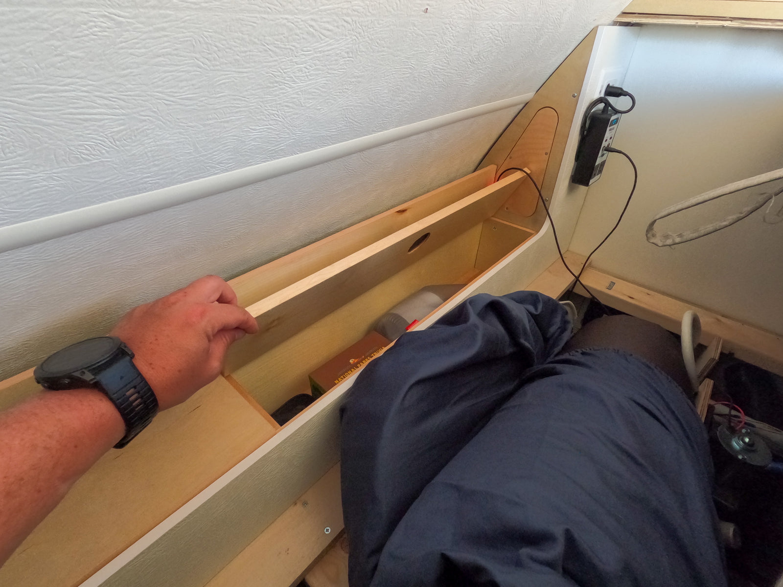 RV rental interior showing a hand with a smartwatch reaching into built-in wooden storage, alongside a sleeping area and power inverter.