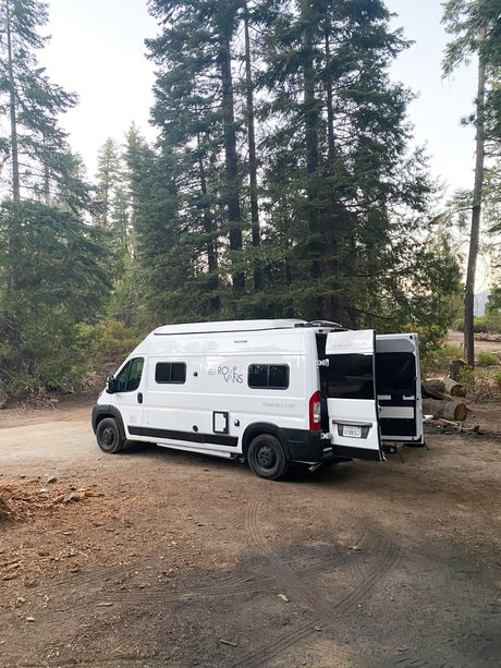 White Rove Vans RV rental parked on a forest dirt road with open rear doors, ready for nature adventure.