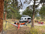 A family relaxes in a hammock, strung between pine trees, next to their RV rental campervan at a serene campsite.