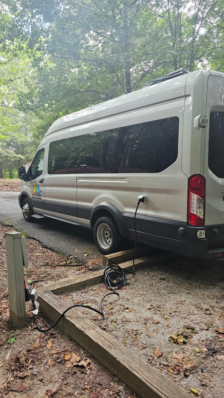 A light-colored WanderVans rv rental camper van is parked, plugged into an electrical hookup at a rustic campsite.