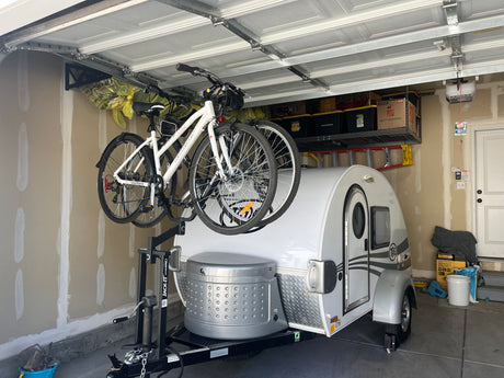 Small white RV (teardrop camper) with bikes on its front rack, neatly stored in a garage, perfect for an rv rental.