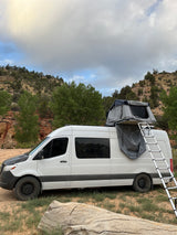 A white Sprinter RV rental with a rooftop tent and ladder deployed, parked for camping in a rocky, tree-lined landscape under cloudy skies.