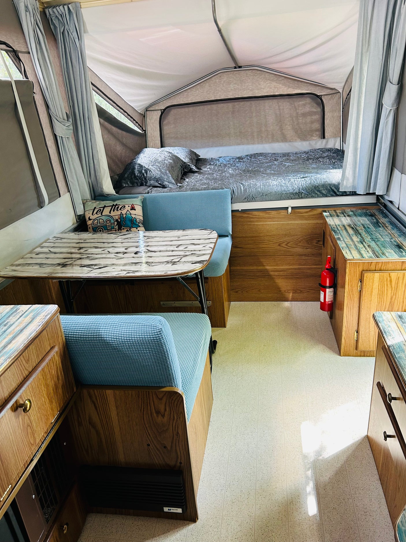An RV rental interior featuring a gray bed, dining table, blue cushioned benches, and light wood-tone cabinetry. A fire extinguisher is mounted.