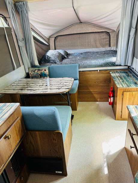 An RV rental interior featuring a gray bed, dining table, blue cushioned benches, and light wood-tone cabinetry. A fire extinguisher is mounted.