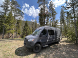 Grey Mercedes Sprinter camper van (RV rental) parked in a sunny forest clearing surrounded by tall pine trees.