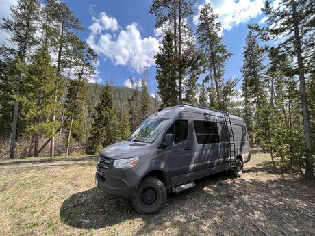 Grey Mercedes Sprinter camper van (RV rental) parked in a sunny forest clearing surrounded by tall pine trees.