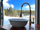 RV rental kitchen setup: a brass faucet and white enamel bowl on a wooden counter, overlooking a snowy forest and blue sky.