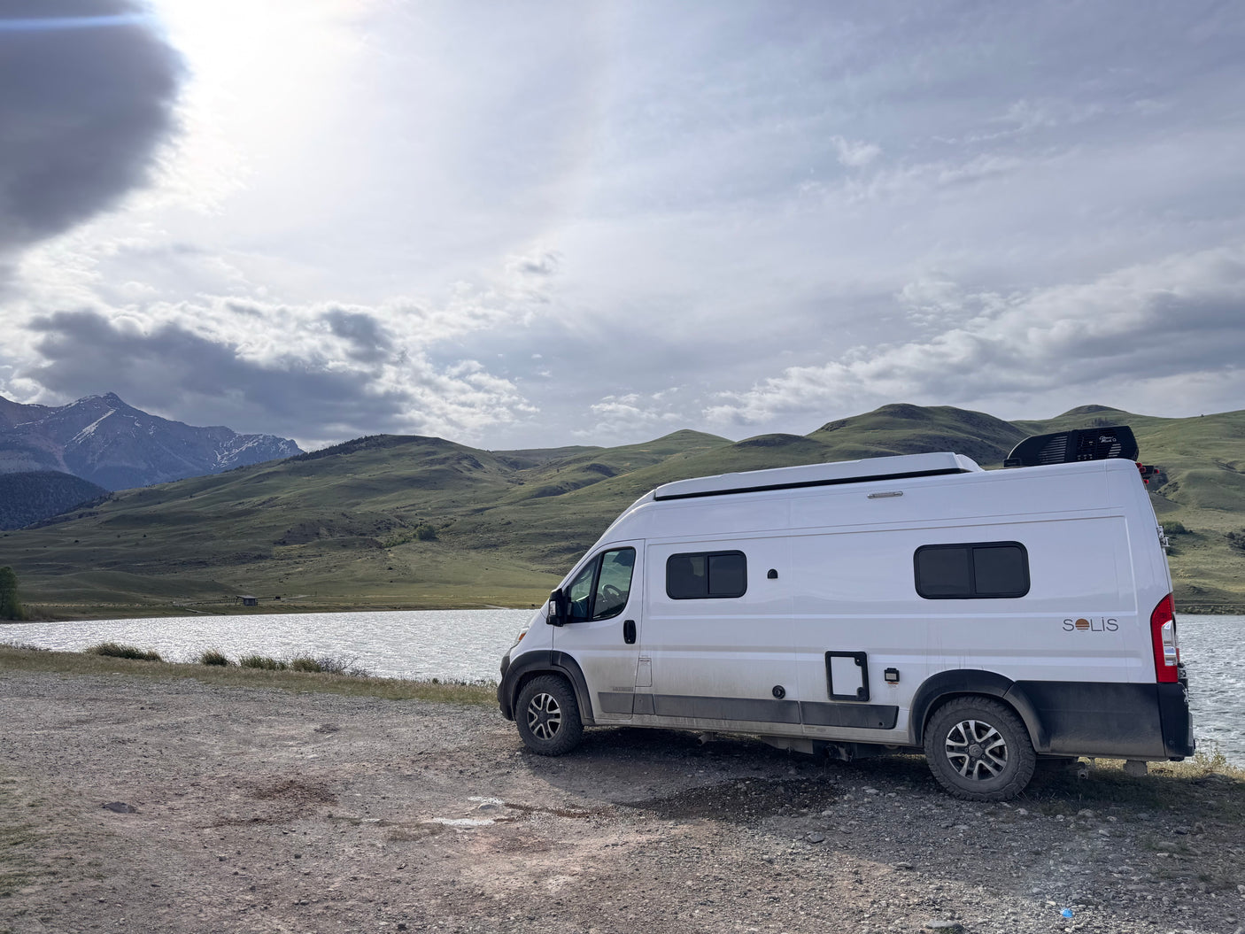 White Winnebago Solis rv rental parked lakeside, offering stunning mountain views under a cloudy sky. Explore the outdoors!
