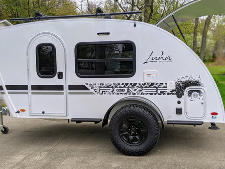 A sleek white Luna Rover teardrop RV rental, featuring a roof rack, large off-road tire, and side amenities, parked on concrete.