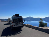RV rental with a bike rack overlooking a stunning, deep blue caldera lake and mountain scenery under a clear sky.