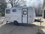 White Scamp compact travel trailer parked on a driveway, ready for rv rental adventures. Features include roof AC, propane tank.