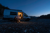 A person sits by a glowing campfire next to an RV rental camper van on a rocky riverbed at dusk, ideal for adventure.