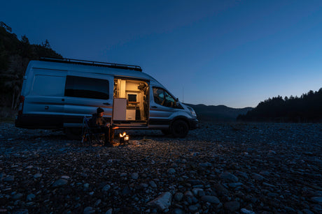 A person sits by a glowing campfire next to an RV rental camper van on a rocky riverbed at dusk, ideal for adventure.