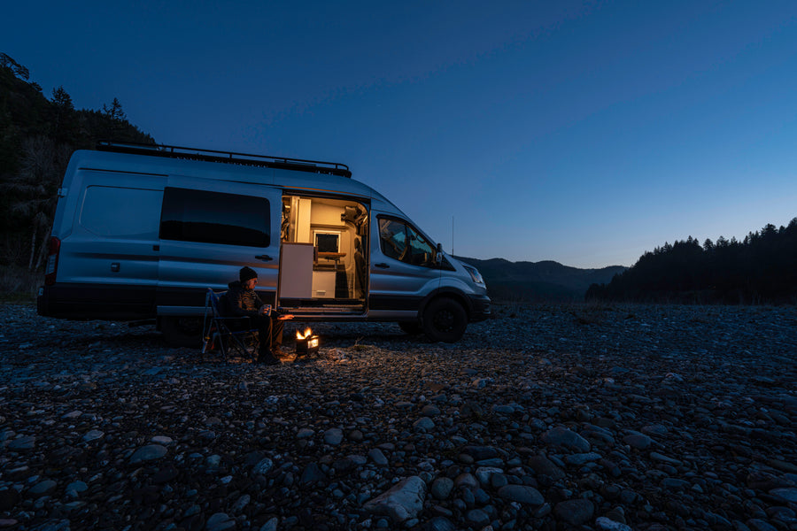 A person sits by a glowing campfire next to an RV rental camper van on a rocky riverbed at dusk, ideal for adventure.