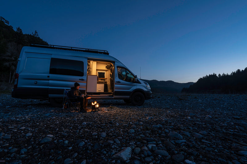 A person sits by a glowing campfire next to an RV rental camper van on a rocky riverbed at dusk, ideal for adventure. — photo 19
