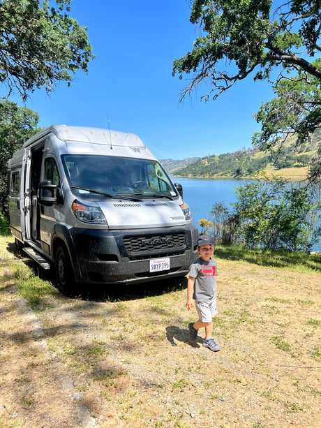 A young boy stands by a silver Winnebago rv rental next to a scenic lake and green hills on a sunny day.