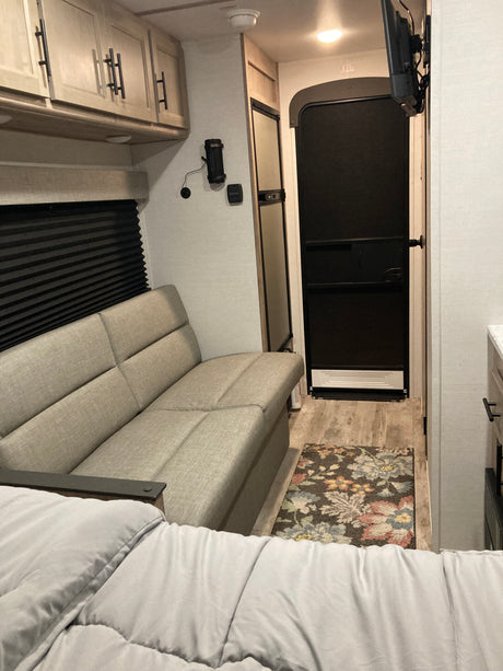 RV rental interior: light wood cabinets, grey sofa, window with blinds, TV, and door in a travel trailer living space.