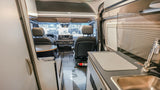 RV rental interior: cockpit, kitchen counter with sink, and seating. View from rear for travel comfort.