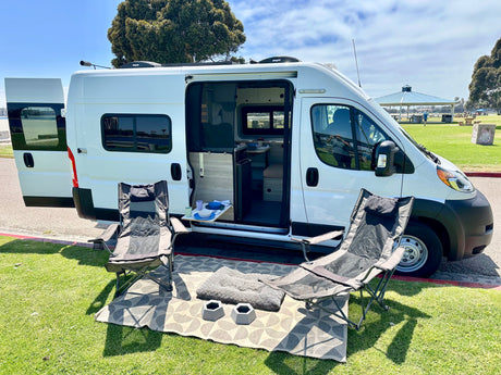 RV rental camper van with open doors showing interior and a relaxing outdoor setup with chairs & pet bowls in a park.