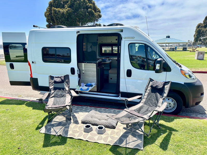 RV rental camper van with open doors showing interior and a relaxing outdoor setup with chairs & pet bowls in a park. — photo 2