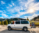Joyful woman in an RV rental van with bikes, parked amidst sunny mountains & pine forests under blue skies.