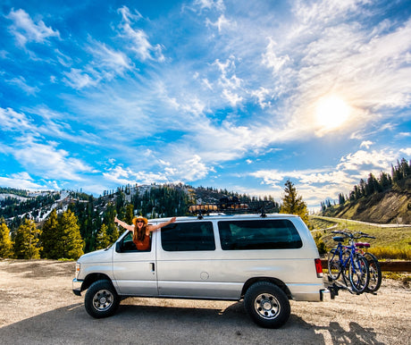 Joyful woman in an RV rental van with bikes, parked amidst sunny mountains & pine forests under blue skies.