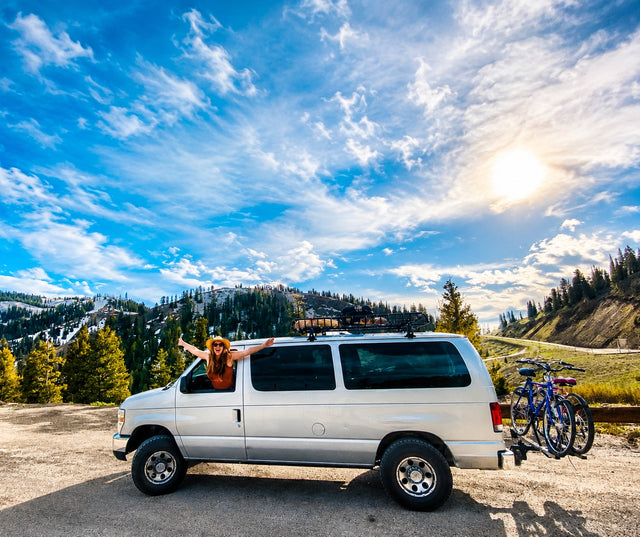 Joyful woman in an RV rental van with bikes, parked amidst sunny mountains & pine forests under blue skies.