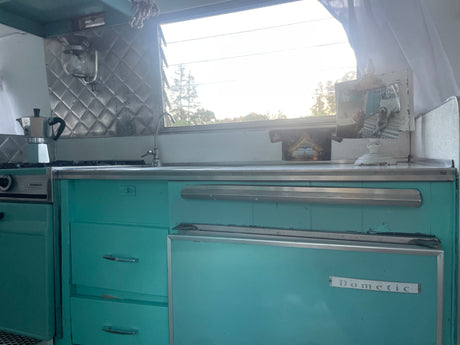 Vintage RV rental kitchen with teal cabinets, Magic Chef stove, Dometic fridge, and moka pot. Bright window view.