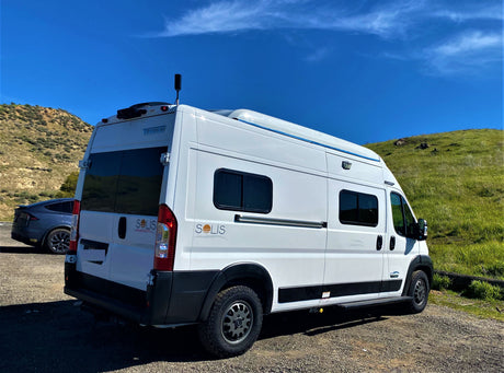 White Winnebago Solis camper van, great for an RV rental, parked on gravel by green hills under a bright blue sky. A Tesla is seen.
