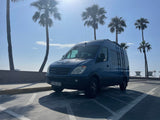 Blue Mercedes-Benz Sprinter camper van (RV rental) parked in a sunny coastal lot with palm trees & lifeguard towers.