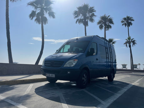 Blue Mercedes-Benz Sprinter camper van (RV rental) parked in a sunny coastal lot with palm trees & lifeguard towers.