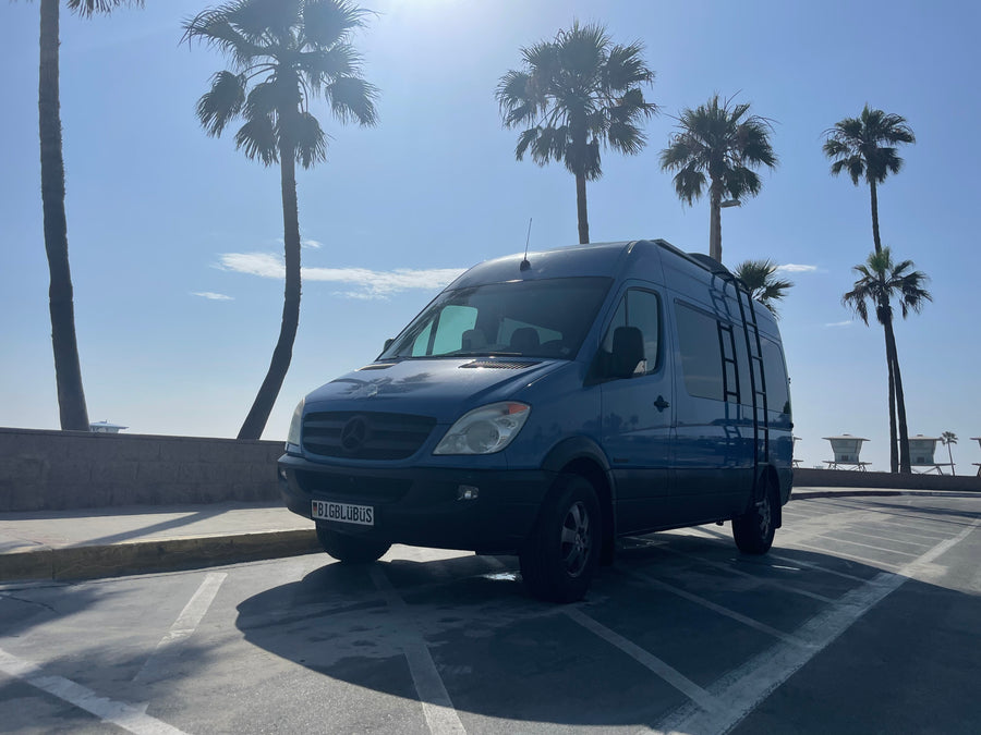 Blue Mercedes-Benz Sprinter camper van (RV rental) parked in a sunny coastal lot with palm trees & lifeguard towers.