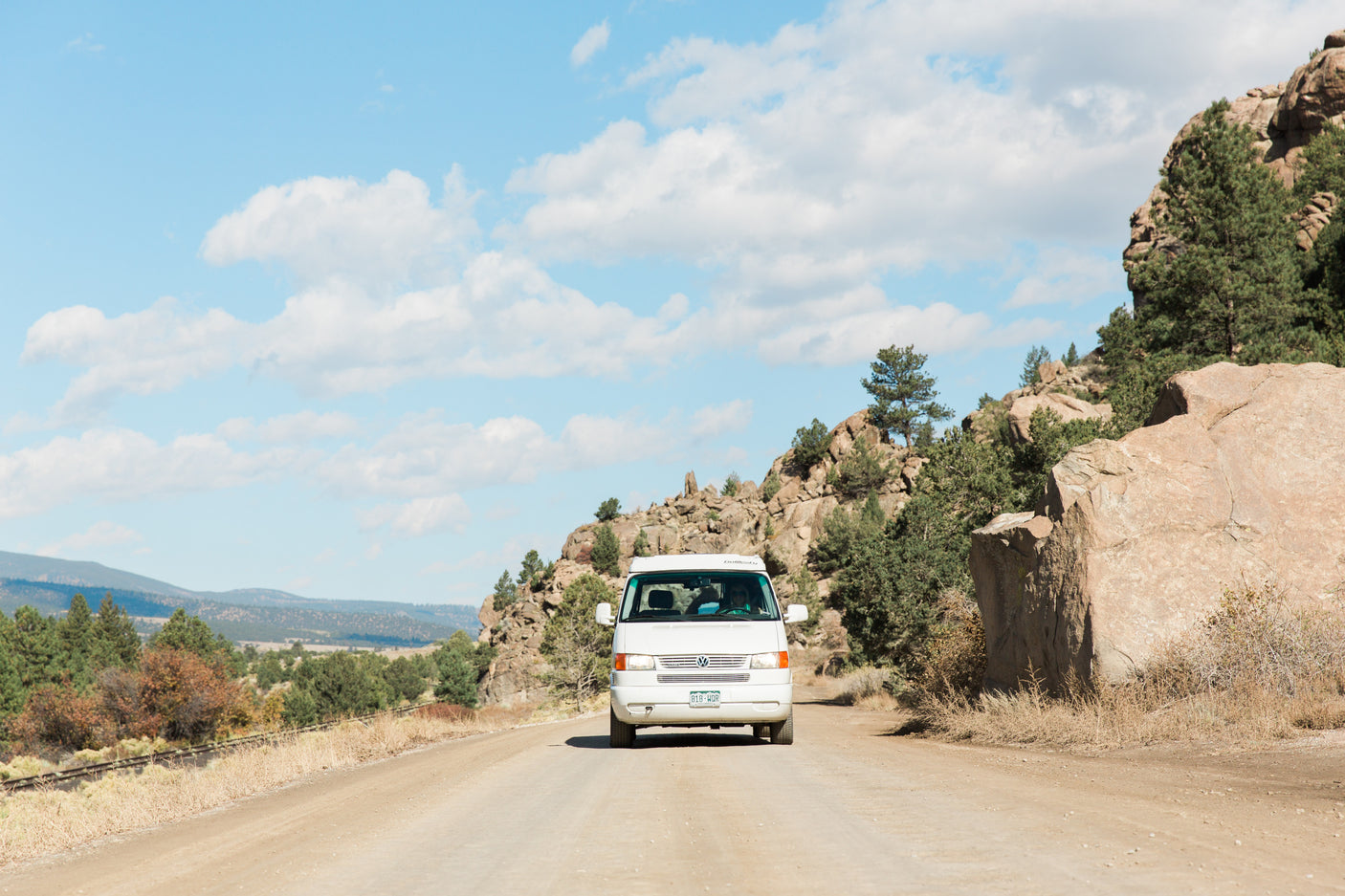 White RV rental campervan on a dirt road, driving through scenic mountains and large rocks under a clear sky.