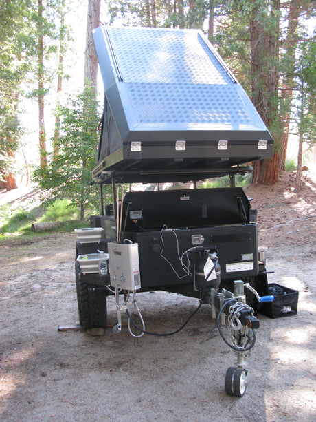 This off-road RV rental trailer features a rooftop tent, solar panel, and external water heater, set up for wilderness camping.