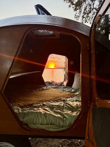 Cozy RV rental view: a bed inside a teardrop camper looking out at a brilliant lake sunset over the water.