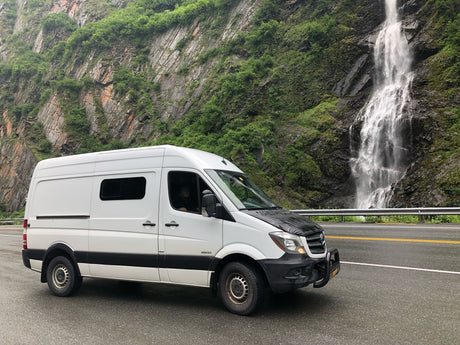 White Sprinter rv rental van beside a majestic waterfall and lush mountain on a rainy day.