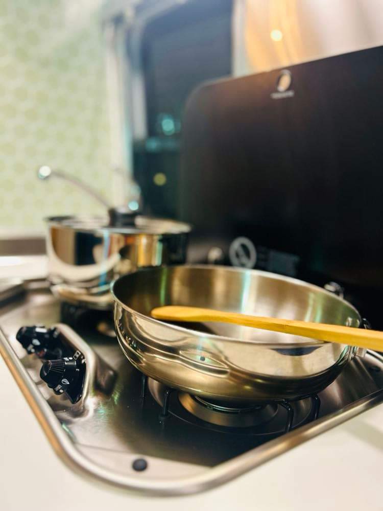 An RV rental's compact kitchen: stainless steel pan with a wooden spoon and a pot on the gas stove, ready for cooking.