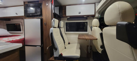 This RV rental interior features a compact kitchen with fridge/microwave, a bed, and cream/grey seating with a dining table.