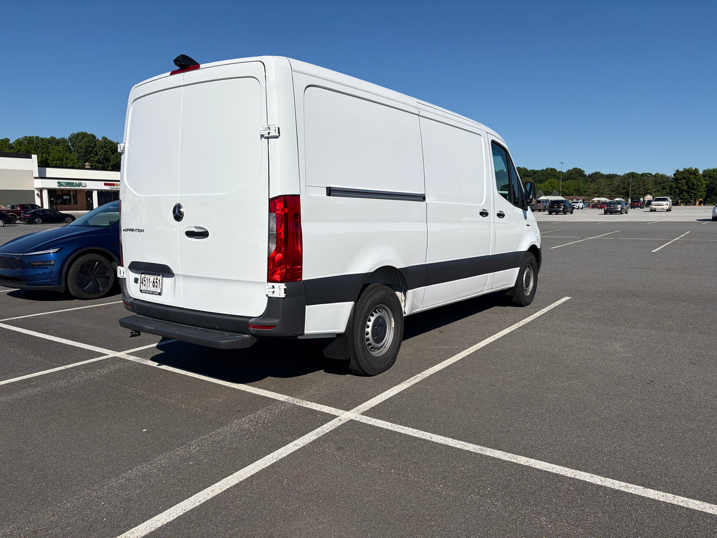 A white Mercedes-Benz Sprinter van, often seen as an rv rental, parked in a sunny lot next to a blue car.