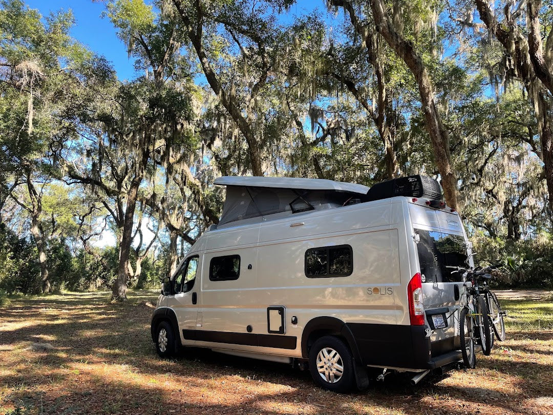 Scenic RV rental: Winnebago Solis camper with pop-top & bikes, parked among iconic moss-draped live oak trees.