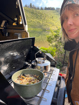 Woman cooking on a camping stove with a kettle by a lake, during an rv rental trip. Nature's beauty enhances the outdoor meal.