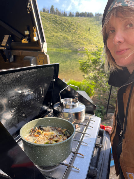 Woman cooking on a camping stove with a kettle by a lake, during an rv rental trip. Nature's beauty enhances the outdoor meal.