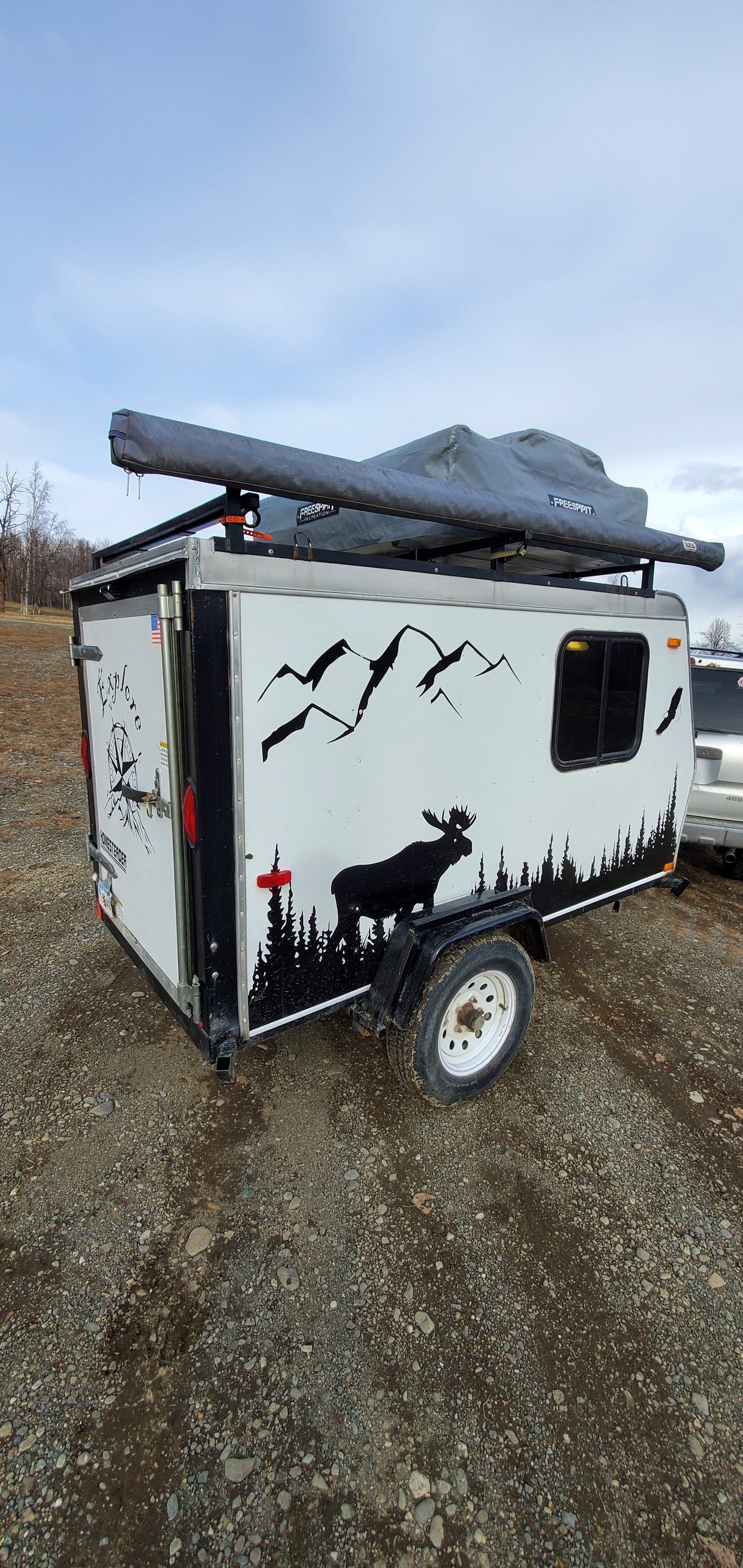 Compact adventure RV rental: A white camping trailer with nature decals and rooftop tent, parked on gravel, perfect for outdoor exploration.