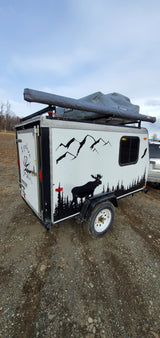 Compact adventure RV rental: A white camping trailer with nature decals and rooftop tent, parked on gravel, perfect for outdoor exploration.