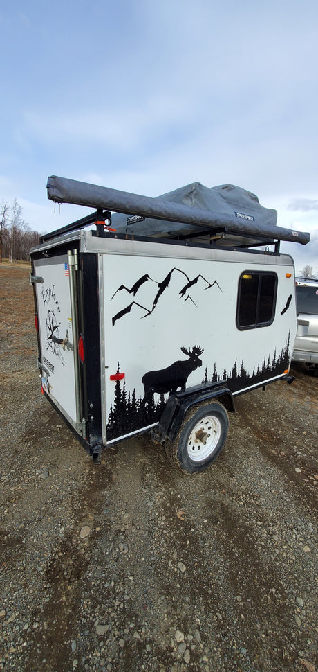 Compact adventure RV rental: A white camping trailer with nature decals and rooftop tent, parked on gravel, perfect for outdoor exploration.